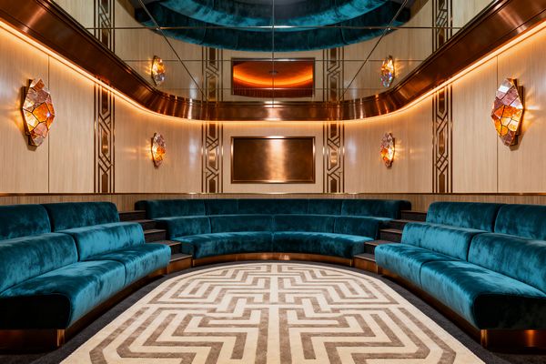 A symmetrical Art Deco media room with tiered, curved teal velvet banquettes facing a bronze-framed screen niche. Blonde wood wall panels carry brass geometric inlays, flanked by faceted amber glass sconces and a continuous warm LED cove. A mirrored ceiling amplifies the glow and height, while dark carpeted steps divide seating. A cream-and-taupe labyrinth-pattern rug centers the space, reinforcing bold geometry and a luxurious theater atmosphere.