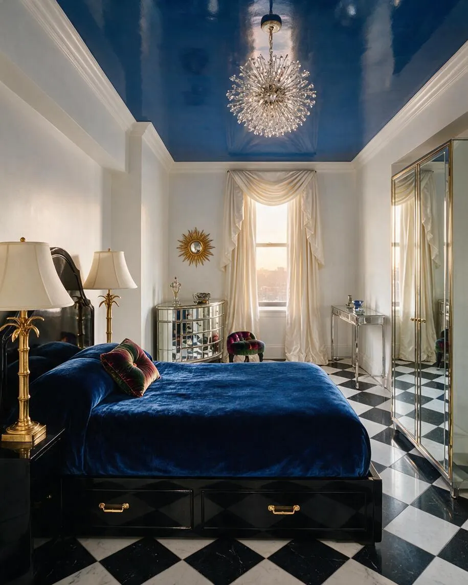 A glamorous guest bedroom features a deep blue velvet bed on a black base, mirrored furniture, and a sparkling starburst chandelier beneath a high glossy blue ceiling. Cream drapery frames the window, while black-and-white diamond tile flooring and gold accents create a dramatic Hollywood Regency look.