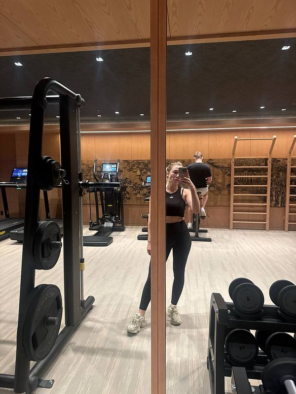 A woman poses in a gym, reflected in a mirror, while another person exercises in the background.