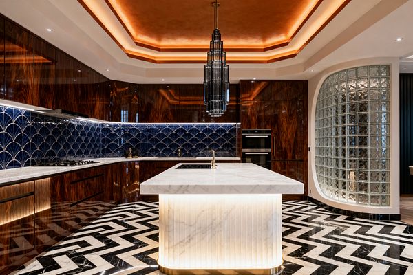 A glamorous Art Deco kitchen features a white marble island with a fluted, backlit base and brass faucet, centered on a bold black-and-white zigzag marble floor. Glossy dark wood cabinetry surrounds a deep navy scallop-pattern tile backsplash with under-cabinet lighting, while a tiered smoked-glass chandelier hangs beneath a layered tray ceiling washed in warm amber light. A curved glass-block wall and integrated black-glass oven complete the sophisticated, streamlined space.