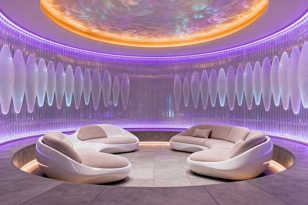 A circular, sunken media lounge features three sculptural, glossy white sofas with taupe fabric cushions arranged around large gray tiles. Curved walls with ribbed translucent panels and frosted ovoid diffusers glow in purple and blue LEDs, while a domed ceiling with a warm amber halo casts an iridescent shimmer, creating an immersive futuristic atmosphere.