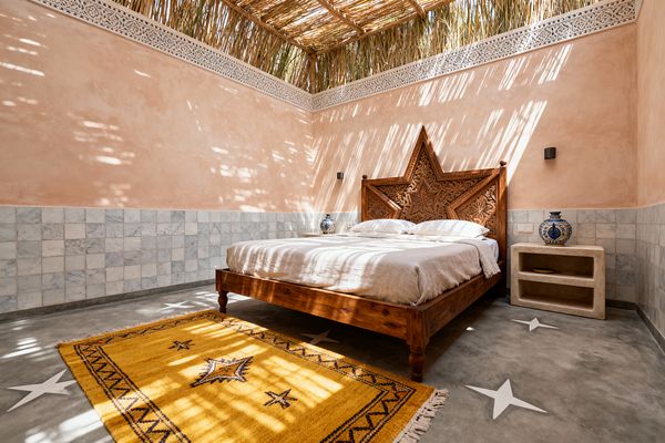 Sunlight filters through a reed ceiling into a Moroccan bedroom with blush plaster walls and pale gray-and-white zellige tile wainscoting. A carved solid-wood bed with a star-shaped headboard stands on a polished concrete floor inlaid with white stars, complemented by neutral linen bedding, a mustard handwoven wool rug, a small tadelakt bedside niche holding a blue-and-white ceramic vase, and slim black wall sconces.
