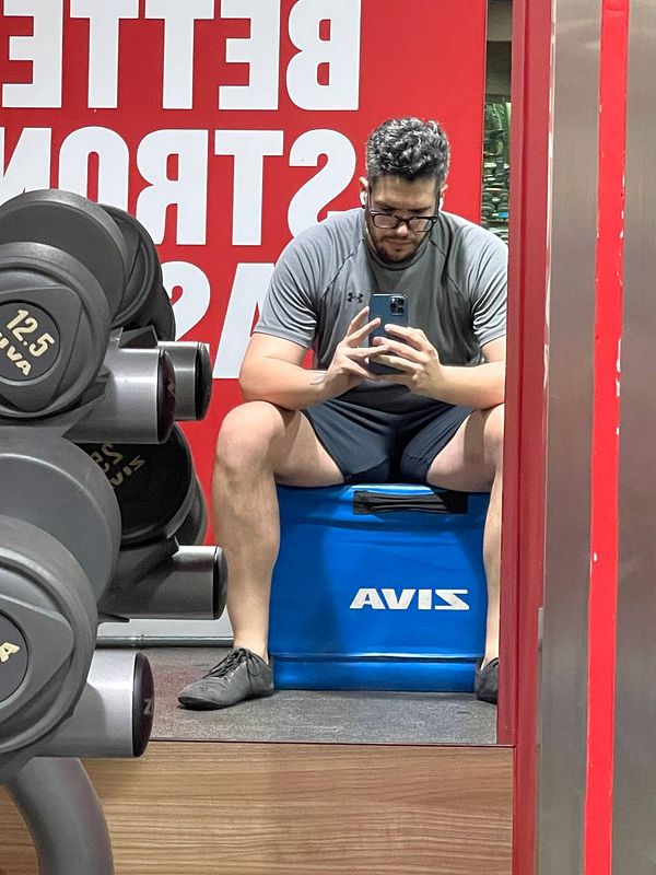 A person is sitting on a blue box in a gym, taking a selfie while surrounded by dumbbells.