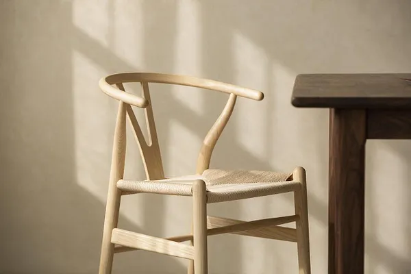 A light wood wishbone-style chair with a woven natural seat sits beside a dark wood table in warm sunlight. The neutral wall behind it casts soft shadows, emphasizing the chair’s sculptural frame and minimalist Japandi aesthetic.