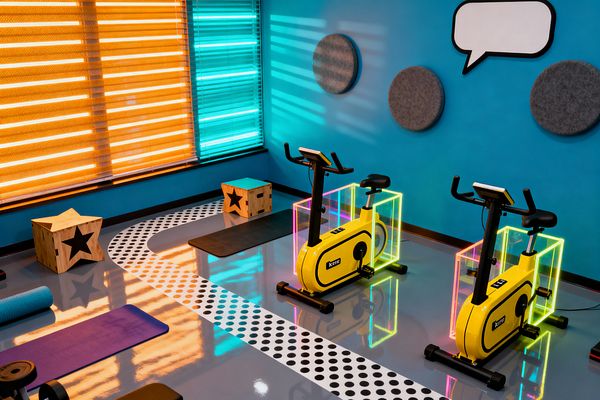 A colorful home gym features two yellow stationary bikes framed by neon-lit clear acrylic boxes on a glossy gray floor. A white path with black polka dots curves between purple and black exercise mats and two plywood boxes with black star cutouts. Large windows have bright orange and teal striped blinds that cast colored reflections. A cyan wall holds round gray acoustic panels and a white comic-style speech bubble, creating a playful, graphic, energetic space.