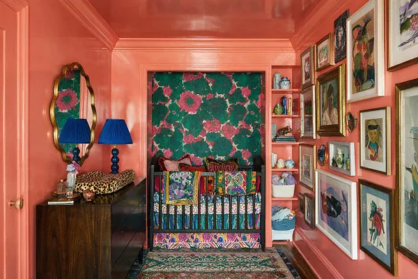 A maximalist nursery with glossy coral walls and ceiling, a dark crib centered in a recessed alcove, and vibrant floral wallpaper behind it. The room is filled with framed artwork, a gold mirror, blue lamps, patterned textiles, and colorful shelves, creating a richly layered, playful space.