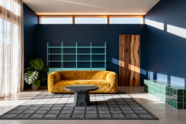 A modern living room with deep navy walls features a low, rounded mustard boucle sofa before a mint-teal open metal shelving unit, a round charcoal stone coffee table on a gray grid-pattern rug, and an emerald green glazed-tile bench to the right. Sunlight enters through clerestory windows, highlighting a warm-toned walnut door, sheer floor-to-ceiling curtains, pale flooring, and a large monstera plant in a concrete pot.