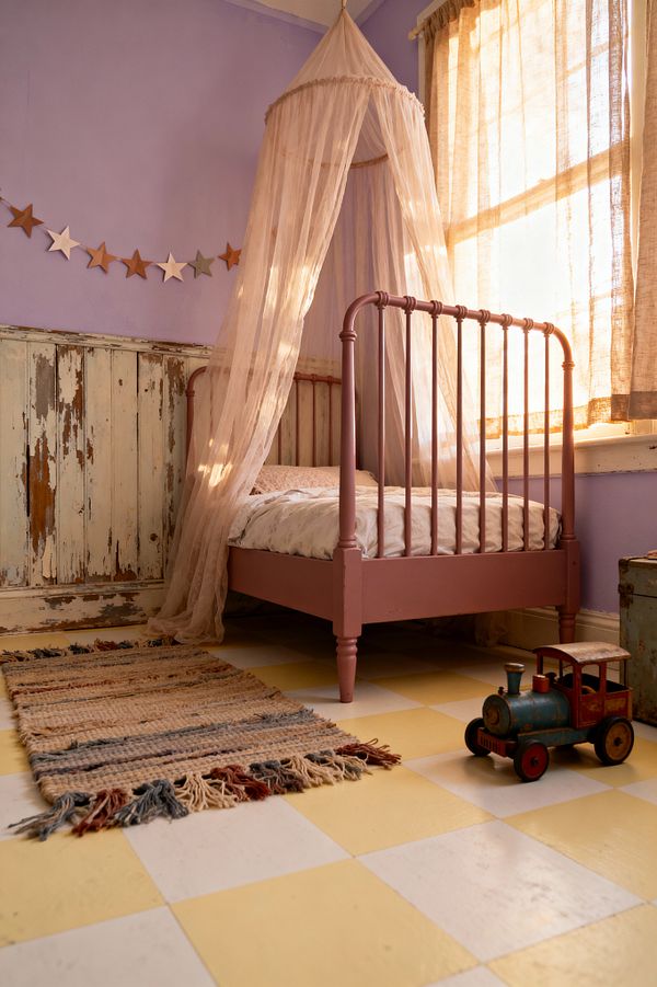 A children’s room with lavender walls and distressed, cream-and-aqua wood wainscoting features a dusty-rose spindle bed draped in a sheer blush canopy. Sunlight filters through beige linen curtains onto a yellow-and-white checkerboard floor with a fringed woven rug. A painted star garland stretches across the wall above the bed, while a vintage metal toy train and a weathered trunk sit nearby, creating a soft, nostalgic atmosphere.