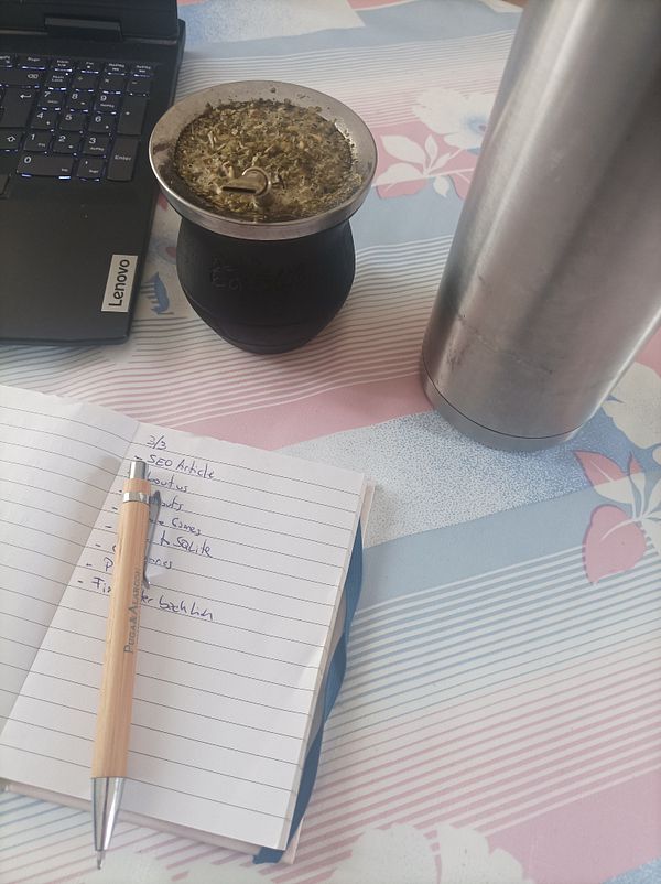 A workspace setup featuring a laptop, a mate cup, a thermos, and a notepad with a to-do list.