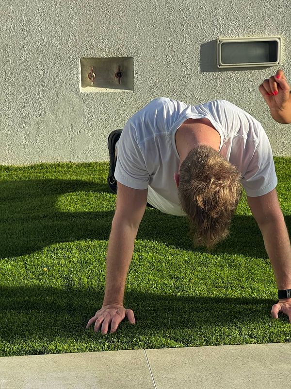 A person is performing push-ups on a grassy surface with a trainer's hand visible nearby.