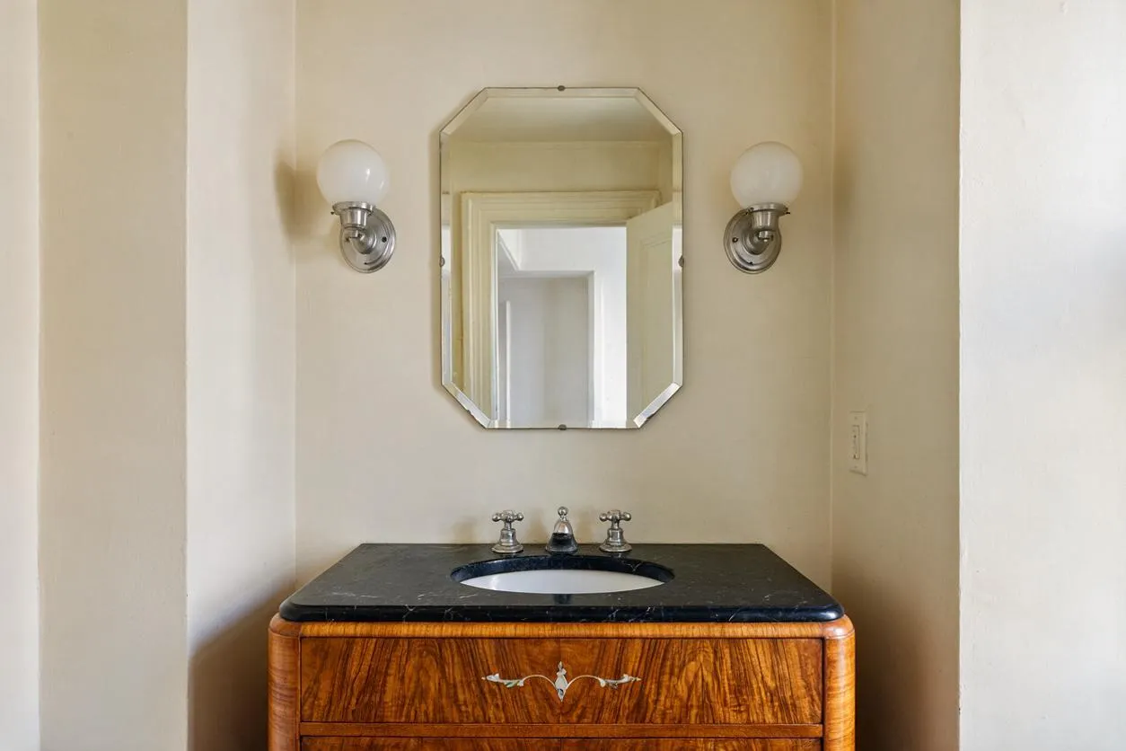 A compact powder room with cream walls, an octagonal framed mirror, and matching globe sconces on either side. Below, a curved wood vanity with a black stone countertop and round sink adds vintage Art Deco character.