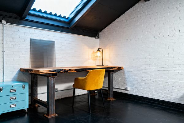 Industrial-style home office under a sloped roof with a large skylight, featuring a thick live-edge wood desk on raw, bolted steel legs against white painted brick with exposed conduit. A mustard yellow upholstered armchair sits beside a black caged table lamp casting warm light over a dark polished concrete floor, while a powder-blue rolling metal drawer unit and a brushed steel wall plate add workshop utility.