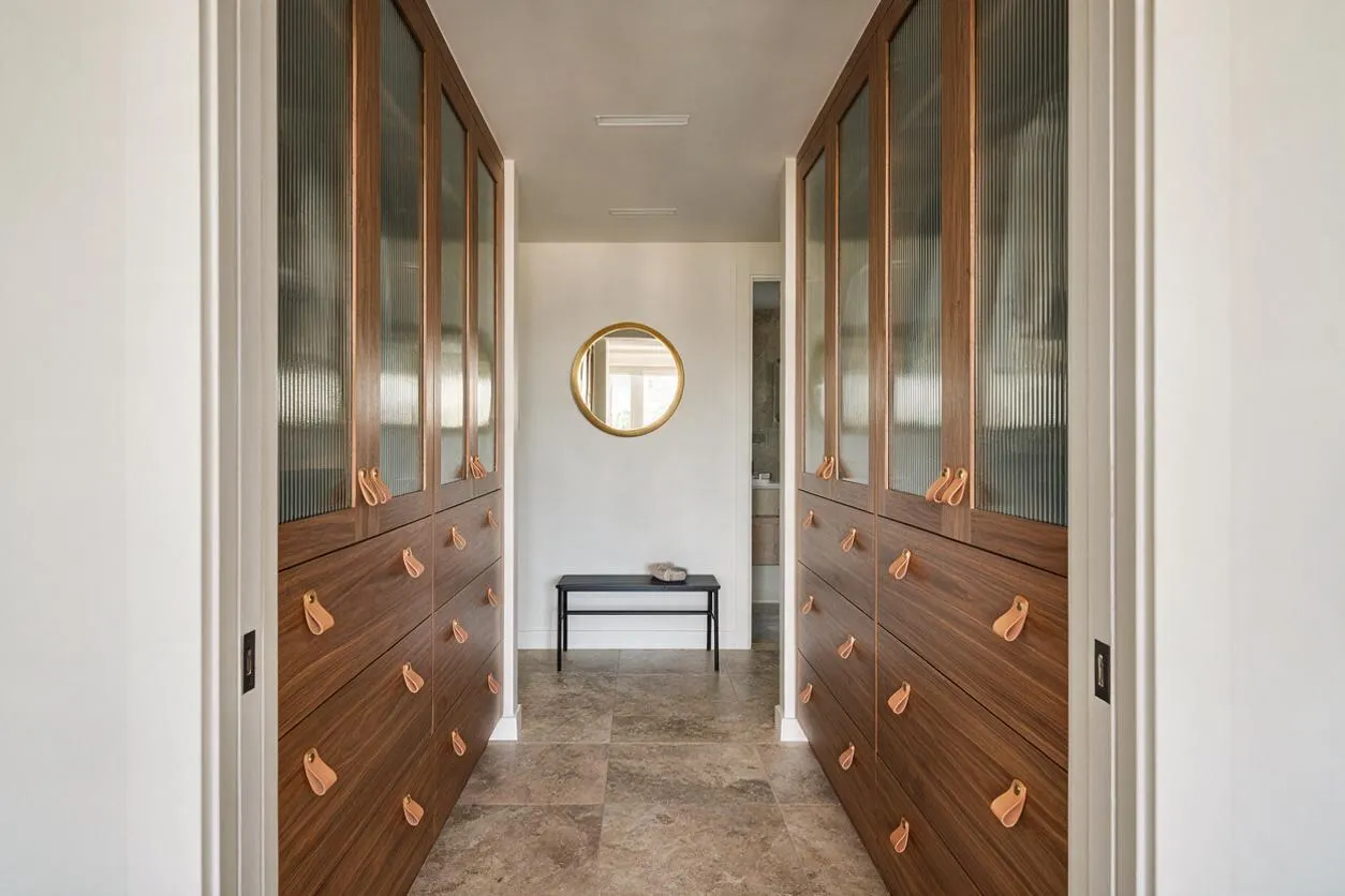 A narrow modern closet hallway with symmetrical built-in wardrobes on both sides, featuring warm wood veneer drawers, copper-toned leather pulls, and upper cabinet doors with reeded glass panels. A round gold-framed mirror and a small black bench sit at the end of the space over large-format stone-look tile flooring.