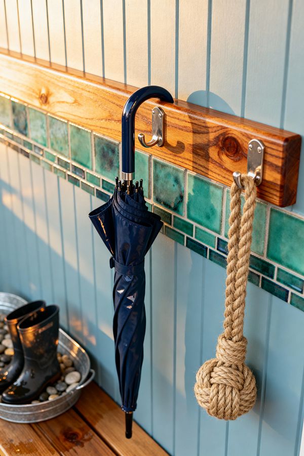 A coastal mudroom detail shows a light aqua beadboard wall with a varnished wood rail and stainless hooks above a band of teal, jade, and deep blue glazed tiles. A navy umbrella and a knotted jute rope hang over warm wood floorboards, while a galvanized metal tub filled with smooth river stones holds wet black rain boots, all lit by low, golden sunlight that highlights the glossy tile and wood grain.