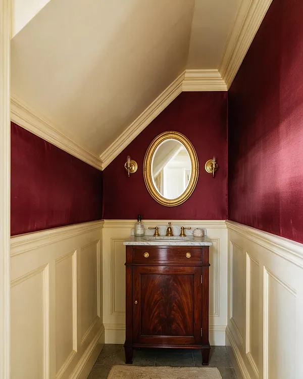 A traditional powder room with deep burgundy upper walls, cream wainscoting, and a sloped ceiling trimmed in matching cream molding. A dark wood vanity with a marble top sits below an oval gold-framed mirror, flanked by two brass wall sconces, creating a refined and formal look.