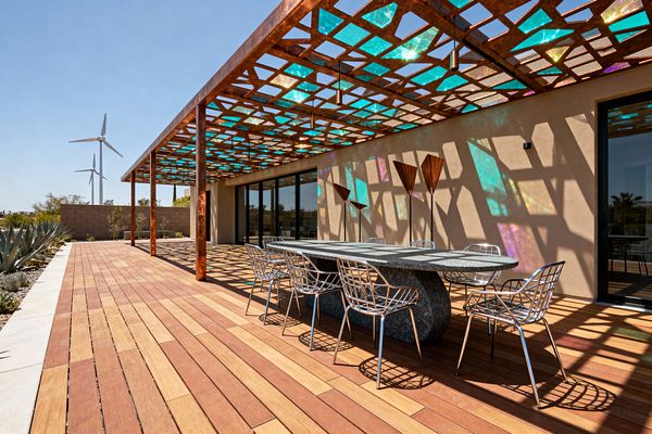 An outdoor patio features a weathered-steel pergola with iridescent dichroic panels that cast colorful geometric shadows across two-tone wood decking and a tan stucco wall. At center sits an oval, honed stone dining table with a sculptural base surrounded by polished metal wire chairs; slim cylindrical pendants hang from the canopy, while black-framed sliding glass doors, desert succulents, and distant wind turbines complete the clean, futuristic scene.