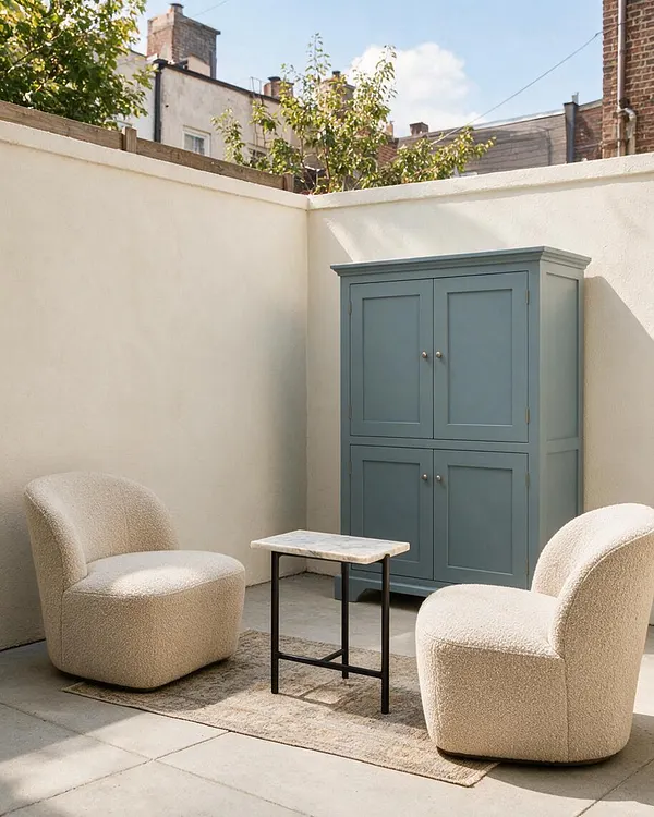 A small transitional backyard seating area with two rounded cream boucle chairs facing a narrow marble-top table. A muted blue storage cabinet sits against cream stucco walls, with a light rug and stone patio flooring under soft daylight.