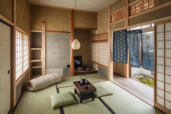 A serene Japanese Zen guest bedroom with tatami mat flooring, low floor cushions, and a small tea table centered in the room. Shoji screens, natural wood framing, and indigo noren curtains create a calm, traditional atmosphere with soft daylight filtering in from the side opening.