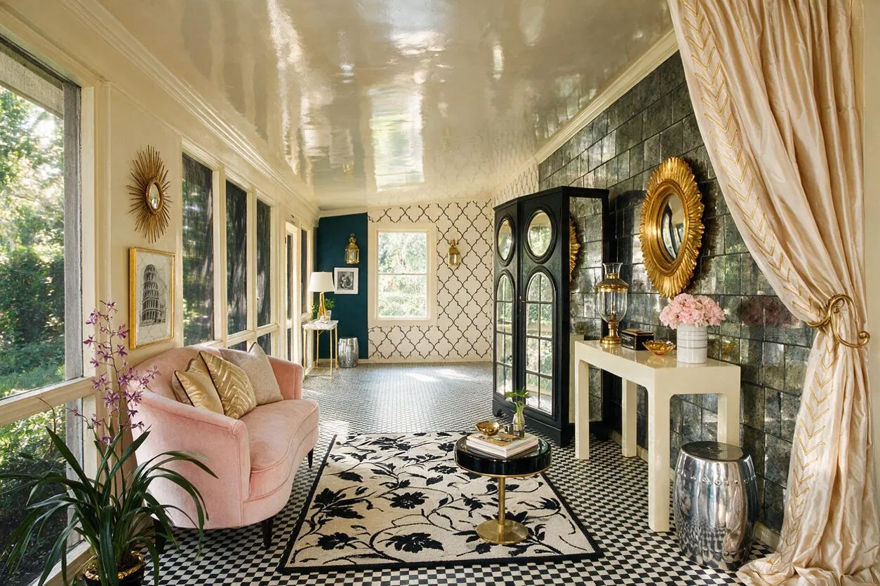 A bright front porch styled like a sunroom, with black-and-white checkered flooring, glossy white ceiling, and mirrored wall decor. A blush pink loveseat, gold accents, patterned wallpaper, a black armoire, and cream drapery create a luxurious Hollywood Regency look.