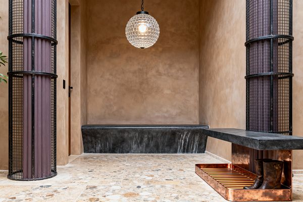 A mudroom with warm tan lime-washed plaster walls and a speckled beige terrazzo floor shows a recessed charcoal concrete bench at the back and a cantilevered bench with a black concrete top over a burnished copper boot tray with a slatted base and a pair of boots. Two mesh-clad columns frame the space, and a clear glass globe pendant hangs centrally, casting soft, warm light.