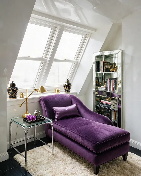 A bright sunroom with slanted skylight windows, white walls, and a purple velvet chaise lounge centered on a cream shag rug. A mirrored bookcase, brass lamp, glass side table, and decorative jars add polished Hollywood Regency glamour.