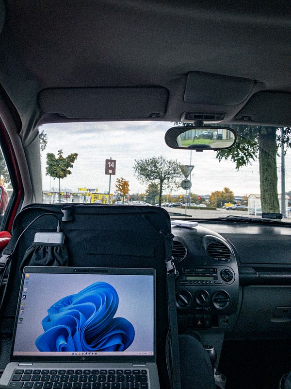 A laptop is set up inside a parked car, with a view of a parking lot and trees outside.
