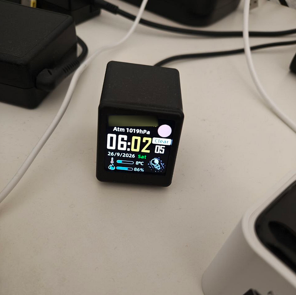 A compact digital clock on a desk displaying the time as 06:02 with weather and atmospheric information.