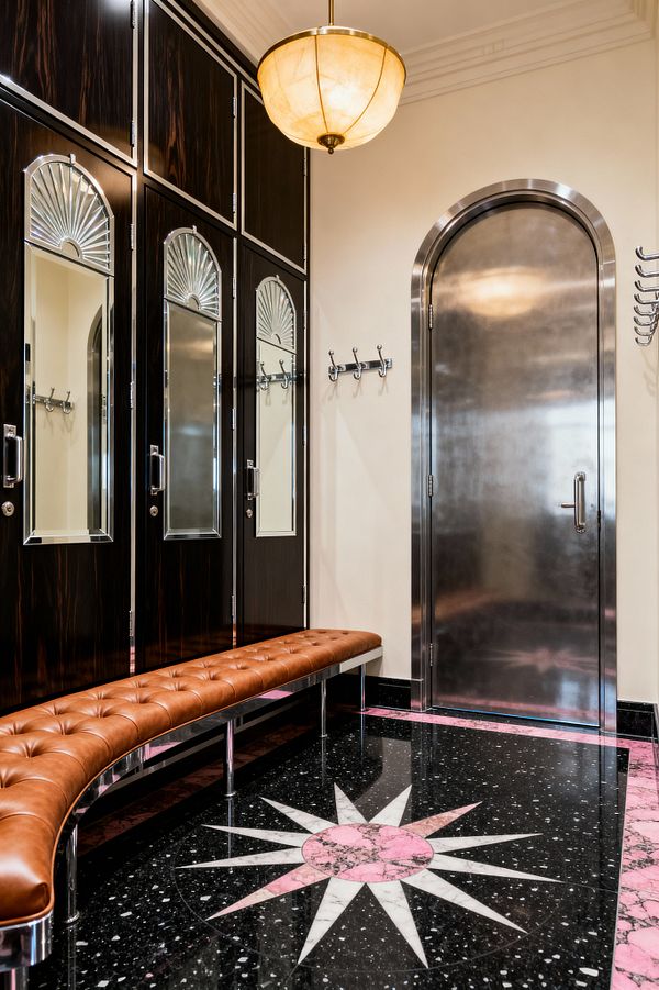 An Art Deco mudroom with a curved, tufted caramel leather bench on chrome legs, set before tall dark wood lockers with mirrored panels and fan motifs. A brushed stainless-steel arched door faces the seating, with polished coat hooks on cream walls. The black terrazzo floor features a pink-and-white starburst medallion and a matching pink border. Overhead, a warm alabaster bowl pendant lights the space.