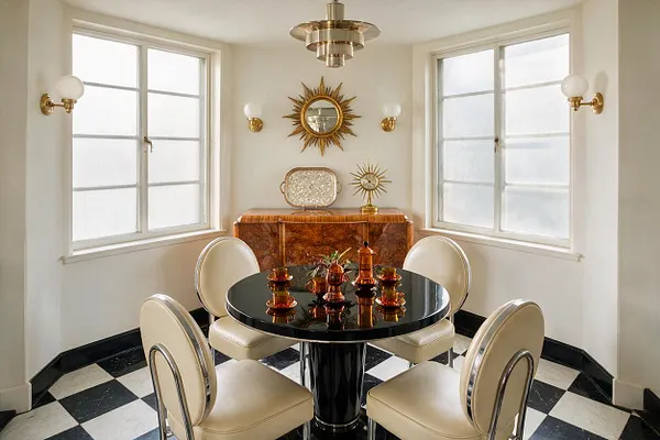 A bright sunroom with four frosted windows, black-and-white checkered flooring, and a glossy black round table surrounded by cream upholstered chairs with chrome trim. The walls feature gold sunburst mirrors and brass sconces, with a sculptural brass-and-glass pendant centered above the table and a lacquered wood cabinet against the back wall.