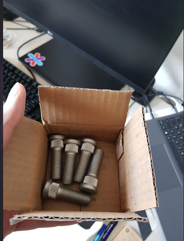 A hand holds a small cardboard box containing several wheel studs.