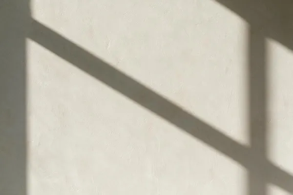 A minimal off-white plaster wall fills the frame, with soft daylight creating strong diagonal and vertical shadows from a nearby window or frame. The matte surface has a subtle hand-finished texture, emphasizing simplicity and natural light.