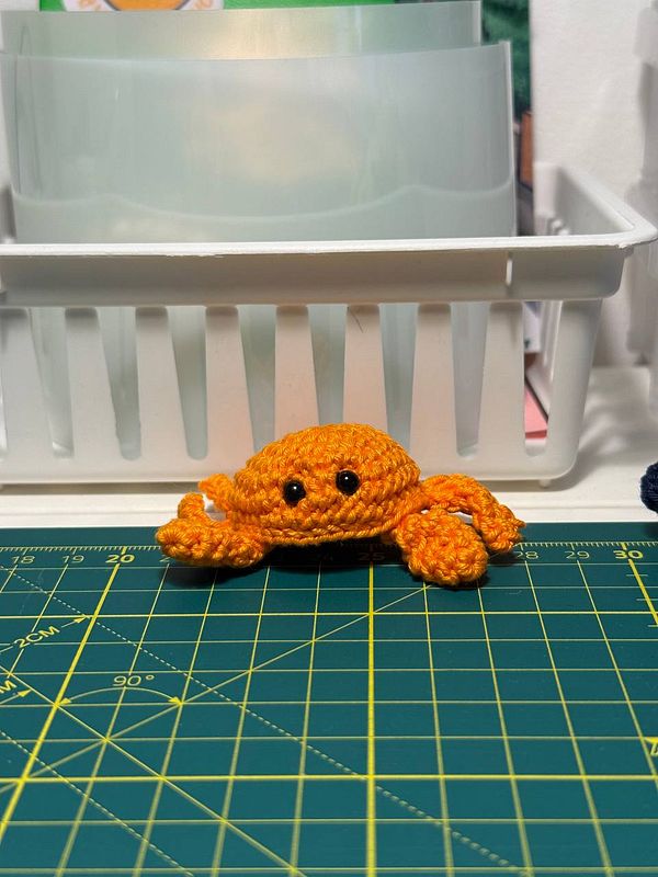 A crocheted orange crab figure is prominently displayed on a green cutting mat.