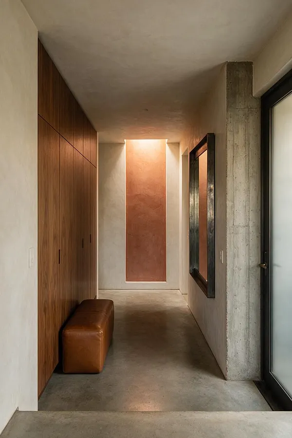 A narrow entryway with smooth concrete floors and walls, a tall wood-paneled storage wall on the left, and a brown leather bench tucked beneath it. A raw concrete column and a black-framed mirror add contrast on the right, while a softly lit terracotta accent wall anchors the end of the hall.