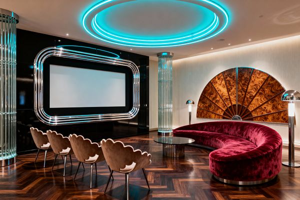 An Art Deco media room with a large screen set in a glossy black wall framed by rounded chrome trim. Aqua neon rings glow in the ceiling and inside fluted ribbed-glass columns. Seating includes a curved burgundy velvet sofa and four scalloped taupe chairs around a black round table on herringbone walnut flooring. A fan-shaped mural in burled wood anchors the right wall, complemented by chrome dome floor lamps.