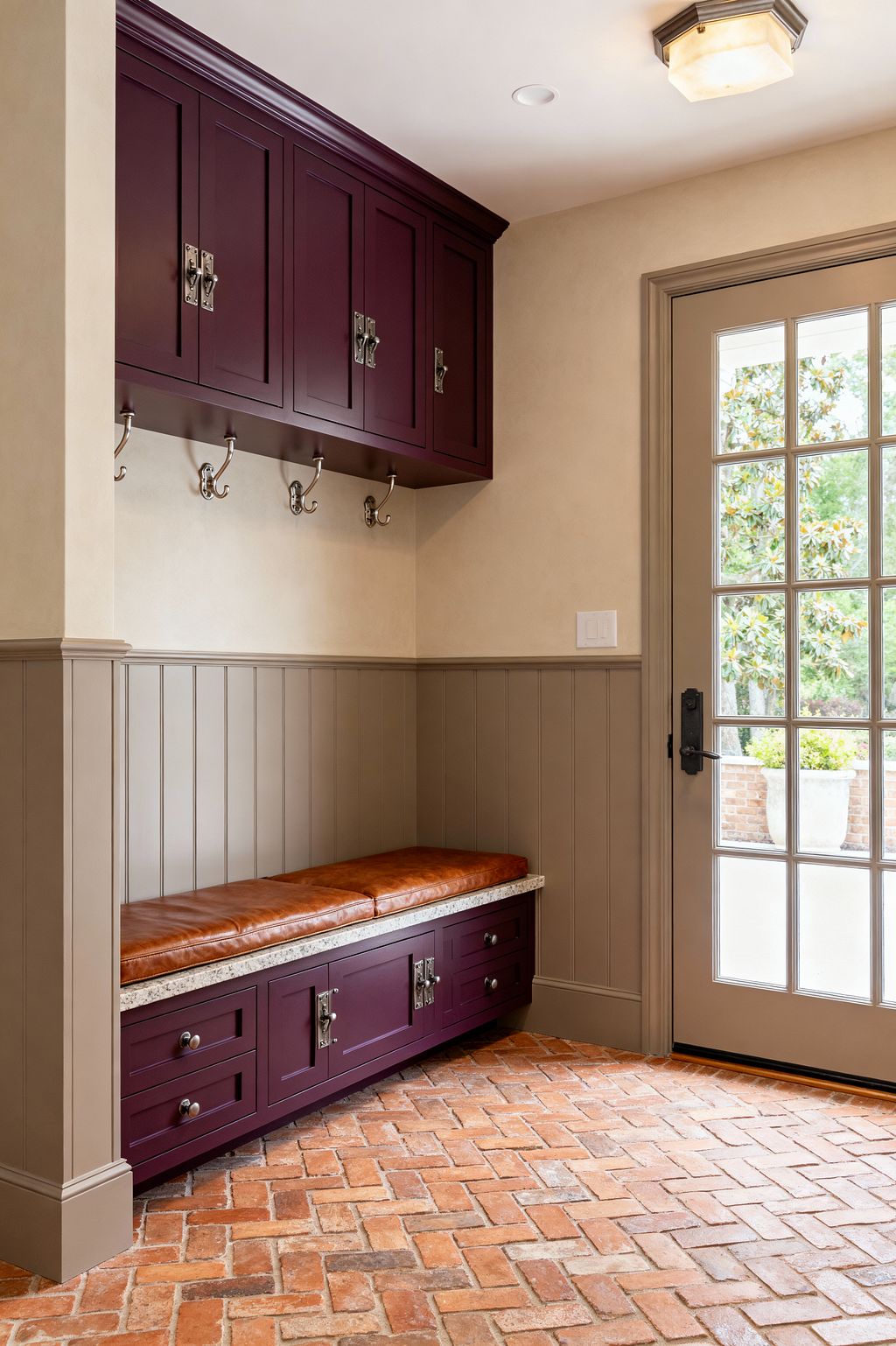A transitional mudroom shows deep aubergine Shaker cabinets above and below a speckled stone bench topped with a cognac leather cushion. Taupe vertical paneling lines the lower walls with polished nickel double hooks beneath the uppers. The floor is herringbone red brick, and a multipane glass door with dark hardware admits daylight. Warm beige walls and a simple square ceiling light complete a functional, refined entry space.