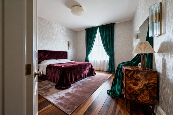 A vintage glam bedroom with a burgundy velvet upholstered headboard and bedspread sits on a plush blush-mauve rug over warm wood floors. Deep emerald velvet curtains frame a tall window with white sheers; silvery textured wallpaper, a glossy walnut burl cabinet with a pleated-shade lamp, ribbed glass sconces with brass details, and a round alabaster-style flush-mount ceiling light complete the softly lit, elegant space.