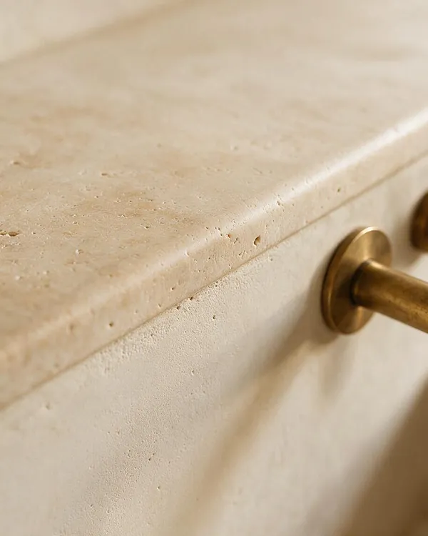 Close-up of a creamy beige stone surface with subtle natural pores and a softly beveled edge, paired with a brushed brass cylindrical handle mounted on the side. The warm neutral tones and tactile materials create a refined Organic Modern detail.