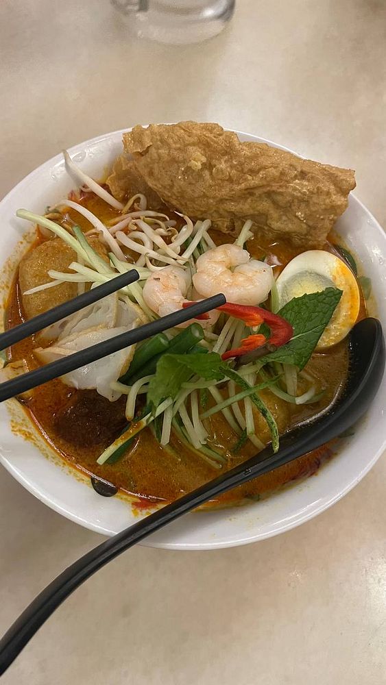 A bowl of curry laksa placed on a wooden mat, accompanied by a spoon and chopsticks, with a condiment bottle visible in the background.