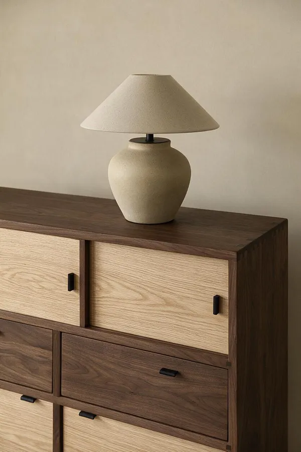 A minimalist Japandi-style table lamp sits on a wooden dresser with mixed light oak and dark walnut drawer fronts. The lamp has a cream ceramic base and a tapered beige shade, complementing the warm neutral wall behind it.