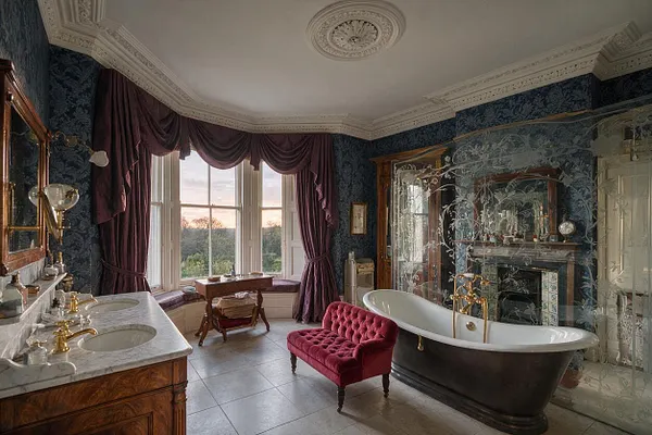 A grand Victorian-style master bathroom with dark patterned wallpaper, ornate white ceiling moldings, and a large bay window dressed in deep plum drapes. The room features a marble-topped double vanity with brass faucets, a dark freestanding clawfoot tub behind a decorative glass screen, and a tufted burgundy bench on pale tiled flooring.