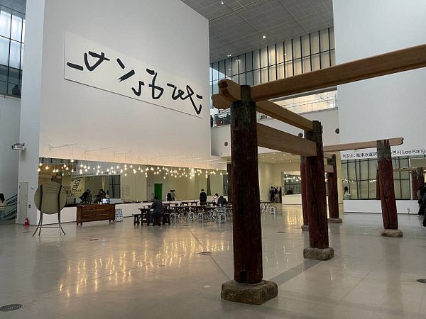 The image captures a spacious art gallery interior featuring a large wall installation and wooden architectural elements.