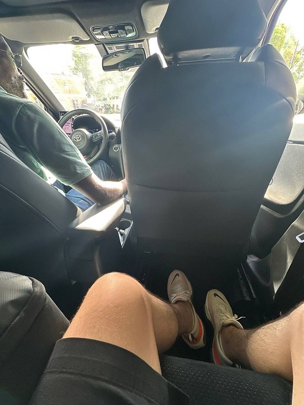 A view from the back seat of a car showing a driver and the passenger's legs.