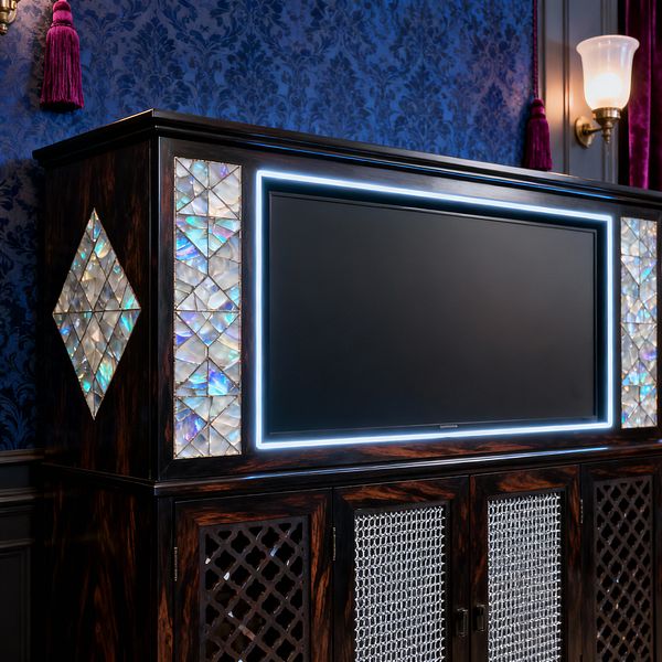 A Victorian-inspired media room shows a dark, high-gloss wood cabinet housing a flat-screen TV outlined by cool LED lighting. Iridescent mother-of-pearl mosaics frame the screen and form diamond motifs, while perforated metal grille doors sit below. The backdrop features cobalt blue damask wallpaper with paneled wainscoting, a brass wall sconce with an opal glass shade, and rich plum velvet with a tassel for a lavish, atmospheric setting.