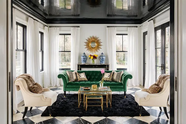 A bright sunroom with white walls and sheer curtains framing tall windows, a glossy black ceiling above, and a black-and-white checkered floor. The room features a deep green velvet sofa, two cream upholstered chairs, a black shag rug, and gold and glass nesting tables with decorative accents.