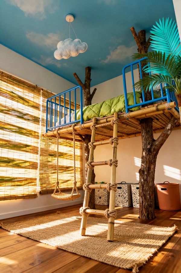 A children's bedroom styled as a tropical treehouse, featuring a bamboo loft bed supported by real tree-trunk posts with bright blue metal rails and a rope ladder. A sky-blue ceiling painted with soft clouds and a cloud-shaped pendant hangs above leaf-green bedding, a small rope swing, woven bamboo blinds, a jute rug, and warm hardwood floors; terrazzo-look storage bins and a terracotta bin line the wall, while a palm frond peeks beside the bed.