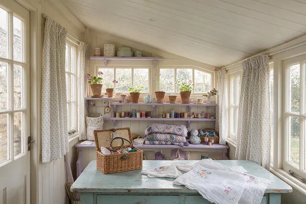 A cozy cottagecore sunroom filled with natural light, featuring many windows with floral curtains, pale lavender shelves lined with potted plants, yarn, and craft supplies. A distressed aqua table in the foreground holds a wicker basket and embroidered linens, creating a soft, handmade, vintage-inspired atmosphere.