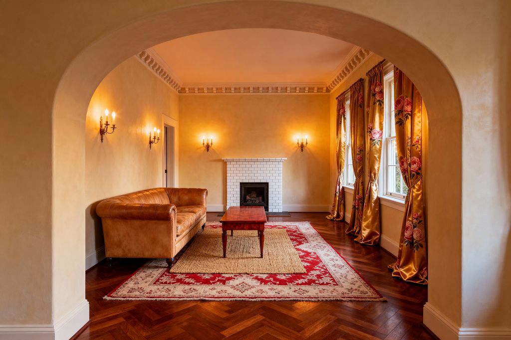 A traditional living room seen through a wide arch features warm plaster walls, dentil crown molding, and a centered fireplace clad in white subway tiles. A caramel leather sofa faces a wood coffee table on layered rugs—an ivory-and-crimson patterned rug over natural jute—while tall windows on the right are dressed in gold satin drapes with large pink roses, and brass candelabra sconces cast warm light over herringbone hardwood floors and white trim.
