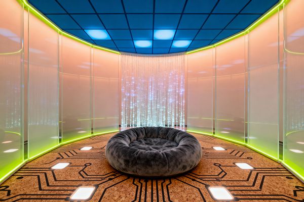 A kids’ room styled like a futuristic pod features curved translucent panels lit by neon-green LED strips, a blue grid ceiling with softly glowing squares, and a cork floor printed with a black circuit-board pattern punctuated by flush square lights. At the center is a large round gray plush nest sofa, while a shimmering fiber‑optic curtain at the far wall casts star-like points of light, bathing the space in a colorful cyberpunk glow.