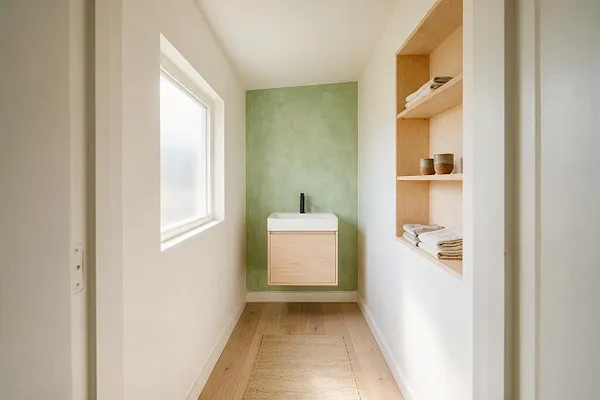 A narrow Scandinavian-style powder room with white walls, a light wood floor, and a frosted window bringing in natural light. At the end of the room is a floating vanity with a white vessel sink and a black faucet set against a soft green accent wall, while built-in wooden shelves on the right hold folded towels and small ceramic bowls.