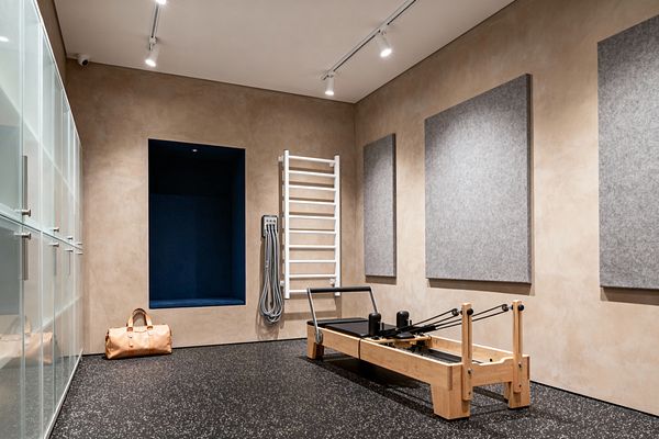 A contemporary home gym with beige troweled plaster walls and black speckled rubber flooring is lit by ceiling track lights. A light oak Pilates reformer stands to the right beside three large gray acoustic panels; a white wall-mounted stall bar and coiled gray cables are nearby. On the left, frosted glass cabinets provide storage, and a deep navy recessed niche sits above a tan duffel bag.